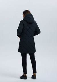 Zwarte gewatteerde pufferjack met capuchon, met een middellange snit en een ruitpatroon. Gedragen met zwarte broek en sportieve schoenen.