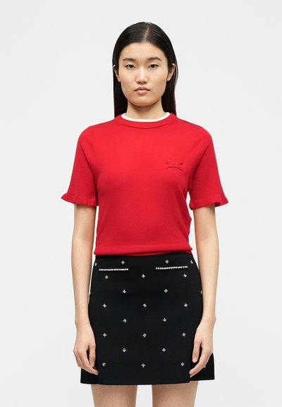 Γυναίκα που φοράει κόκκινο πουλόβερ με κοντά μανίκια και μικρό κεντημένο λογότυπο, και μαύρη μίνι φούστα διακοσμημένη με λευκές πέρλες που μοιάζουν με καρφίτσες.