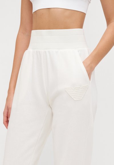 Pantaloni da tuta bianchi con vita alta elastica, tasche laterali e un patch logo testurizzato sulla gamba sinistra. Realizzati in tessuto morbido.