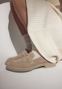 Beige suede loafers with decorative stitching and a small heel, worn with ribbed white socks and a layered cream skirt.