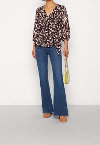 Blumenmuster-Wickeloberteil in Dunkelbraun und Pink mit Ballonärmeln, kombiniert mit ausladenden blauen Jeans. Accessoires sind eine gelbe Handtasche.