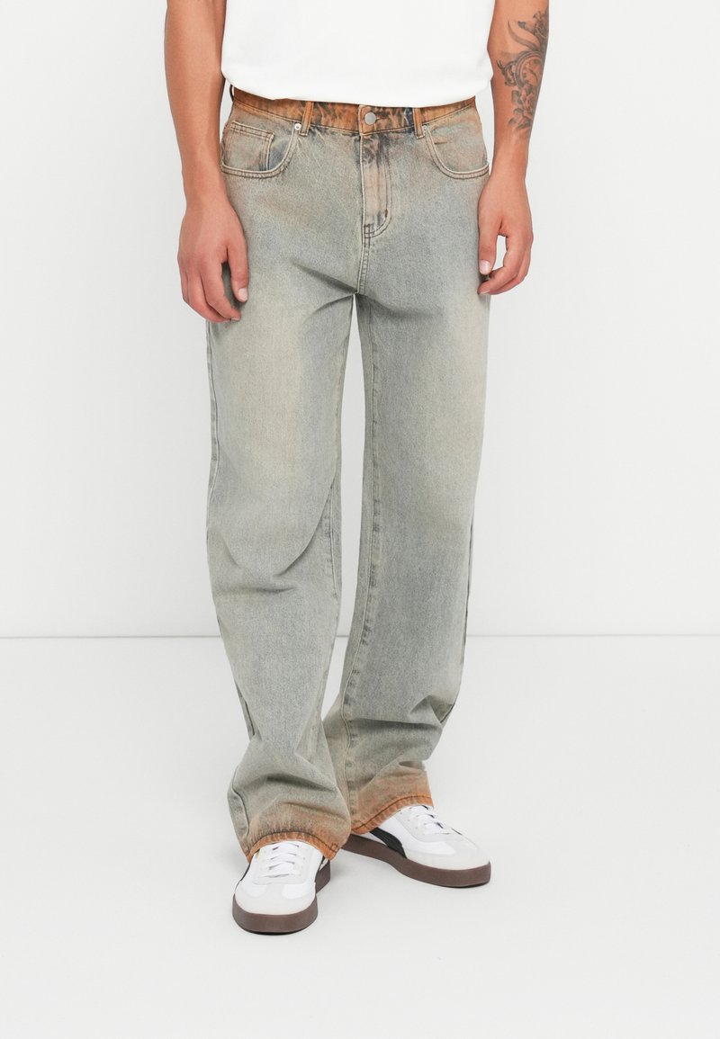 Lichtgrijze denimjeans met een ontspannen pasvorm, met vervaagde plekken en een oranje tint aan de zoom. G gedragen met witte sneakers.