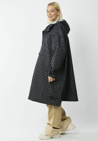 Zwarte regenjas met een rond patroon, voorzien van een capuchon, lange mouwen en een losse pasvorm, gecombineerd met beige broeken en stevige sneakers.