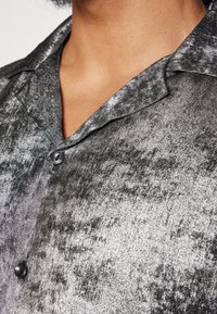 Camicia a maniche corte in tessuto nero metallizzato, con un sottile motivo testurizzato, colletto a punta e chiusura con un singolo bottone nero.