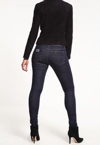 Dunkelblaue Skinny-Jeans mit glatter Textur, ausgestattet mit einer hinteren Gesäßtasche, kombiniert mit einer schwarzen Wildlederjacke und schwarzen hohen Schuhen.