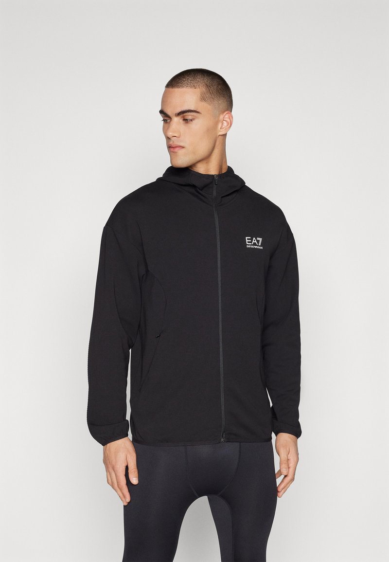 Musta vetoketjullinen takki, jossa on huppu, sivutaskut ja pieni valkoinen logo rinnassa. Valmistettu sileästä, joustavasta kankaasta.