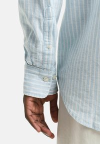 Camicia a maniche lunghe a strisce blu chiaro con bottoni bianchi, caratterizzata da un orlo arrotondato e un tessuto testurizzato. Mano parzialmente visibile.