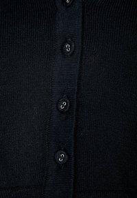 Svart stickad cardigan med sex knäppningar. Texturen är slät med en ribbad mönstring längs kanterna.