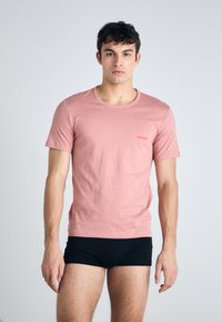 T-shirt rose clair à manches courtes en coton lisse, avec un petit logo rouge "HUGO" sur la poitrine, accompagné de culottes noires ajustées.