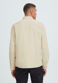 Camisa de manga larga beige hecha de tejido suave, con cuello puntiagudo y puños abotonados, combinada con pantalones oscuros. Vista desde atrás.