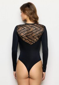 Una donna girata di spalle indossa un body a maniche lunghe nero con pizzo trasparente e motivi a zigzag sulla parte superiore della schiena, su uno sfondo neutro.