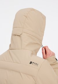 Doudoune beige avec un col montant et une capuche ajustée. Présente un logo sur l'épaule gauche et un tissu texturé avec des sections matelassées.