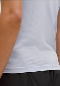 Rechterarm en torso in een gestructureerd lichtgrijs sportshirt en zwarte shorts tegen een effen achtergrond.