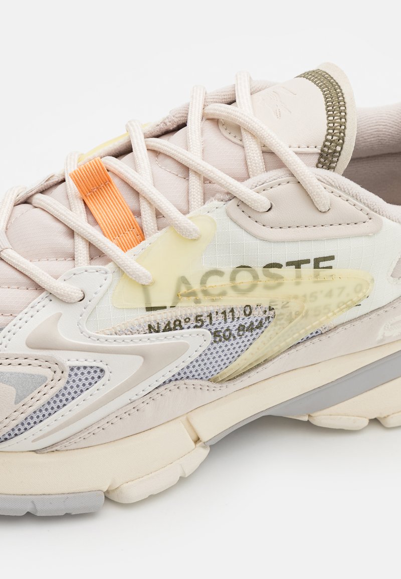 Le sneakers Lacoste presentano una tomaia in tessuto beige chiaro, dettagli in mesh, accenti arancioni e gialli, e una suola bianca curvata con gomma grigia.