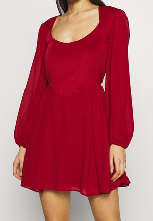 Femme portant une robe rouge à manches longues avec un décolleté en rond et une jupe évasée, visible des épaules jusqu'à la mi-cuisse.