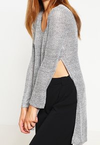 Maglione grigio argento a maniche lunghe con scollatura a V, sfiancato ai lati e in tessuto testurizzato, indossato con pantaloni neri.