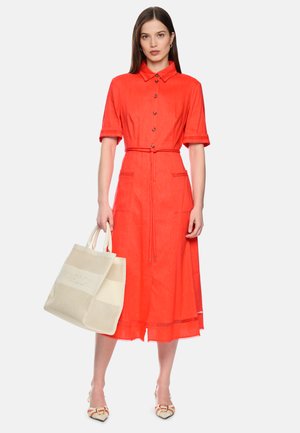 Woman wearing a bright orange midi dress with buttons, short sleeves, and a waist tie, holding a large beige tote bag.