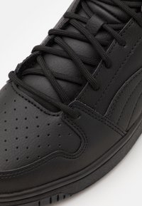 Svart sneaker med en texturerad läderöverdel, perforerad tådel och platta svarta snören. Gummisula och s ömdekor detaljerar designen.