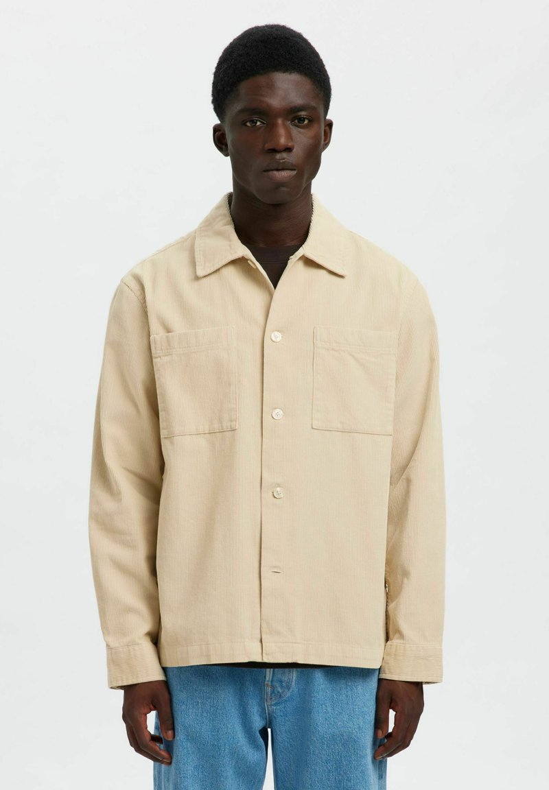 Ung mand med kort hår iført en beige knappet kordjakke og lyseblå jeans, stående mod en ensfarvet baggrund.