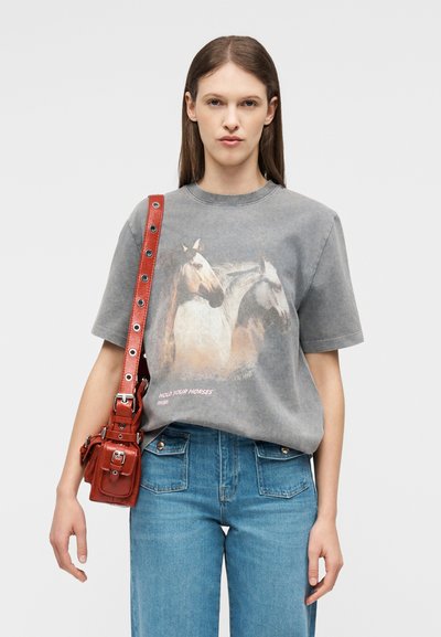 Γυναίκα που φοράει γκρι μπλουζάκι με εικονογραφήσεις αλόγων, μπλε τζιν και κόκκινη δερμάτινη τσάντα ώμου με μεταλλικά φωλιάσματα.
