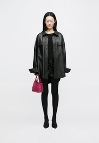 Svart lær oversized skjortejakke, svart topp, blanke sorte shorts, svarte strømpebukser, spisse sko, holder en liten rosa håndveske.