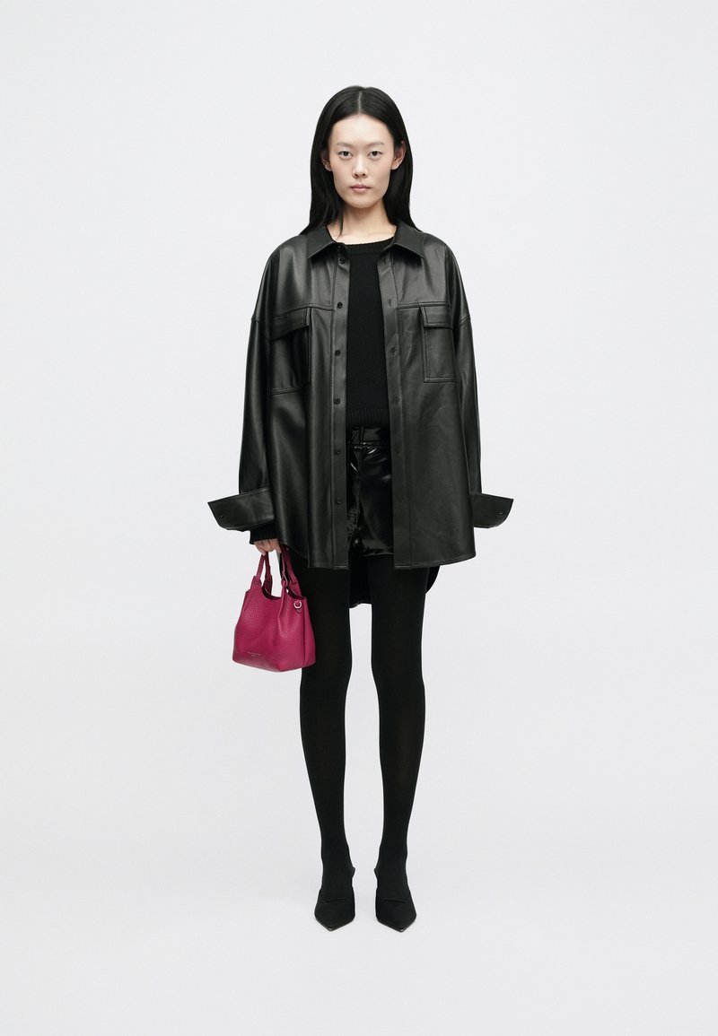 Svart lær oversized skjortejakke, svart topp, blanke sorte shorts, svarte strømpebukser, spisse sko, holder en liten rosa håndveske.