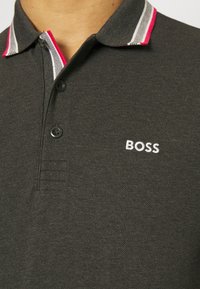 Mörkgrå pikétröja med texturerad tyg, vitt broderat "BOSS"-logotyp och en randig krage med röda, vita och svarta accenter.