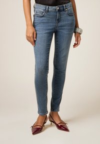 Jeans de mezclilla azul de corte slim con cintura de altura media, ligero desgaste y cinco bolsillos, combinados con tacones de charol burdeos.