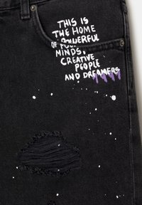 Jean noir avec texte manuscrit blanc et violet indiquant "This is the home of powerful minds, creative people and dreamers" près de la poche.