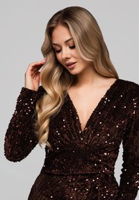 Robe marron à sequins avec un décolleté en V profond et une coupe ajustée. Manches longues et tissu texturé qui reflète la lumière. Coiffure en vagues douces.