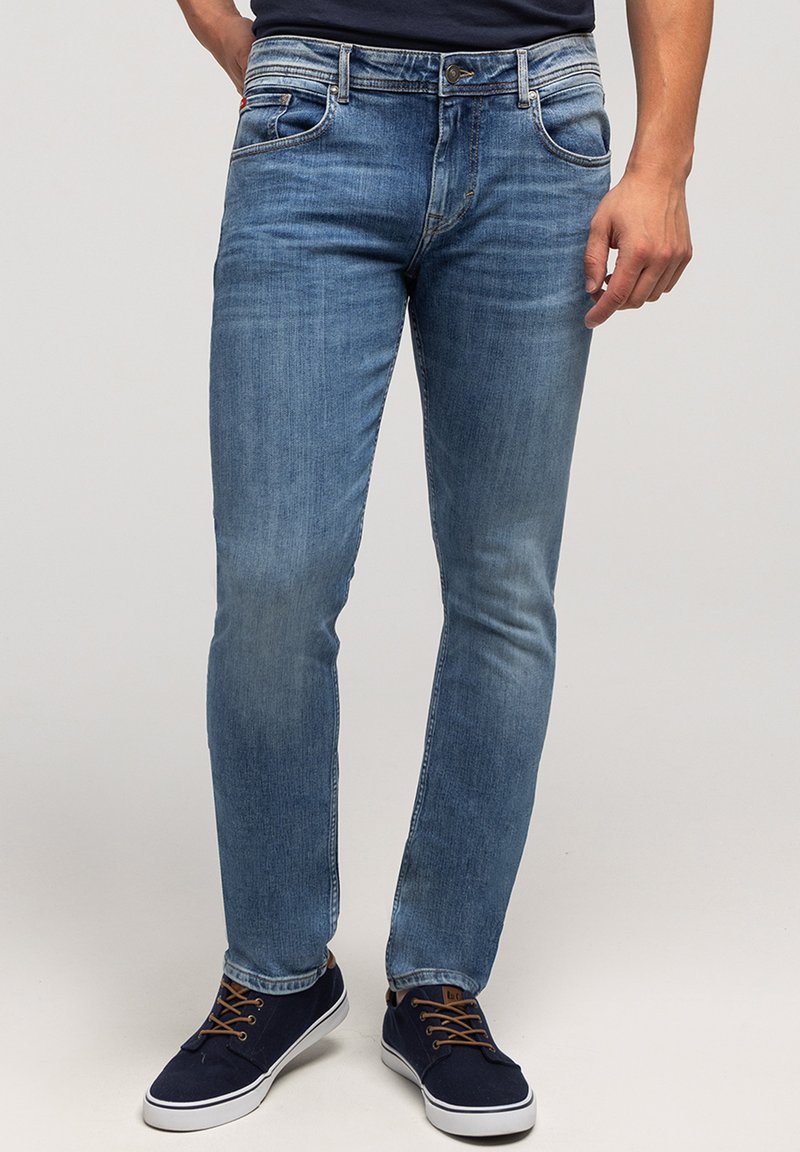 Mężczyzna w dopasowanych niebieskich dżinsach i granatowych sneakersach z brązowymi sznurówkami, stojący na jednolitym jasnym tle.