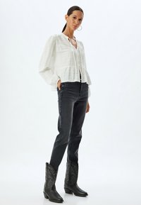 Blusa blanca texturizada con dobladillo acampanado, combinada con jeans ajustados oscuros y botas de cowboy negras con costuras intrincadas.