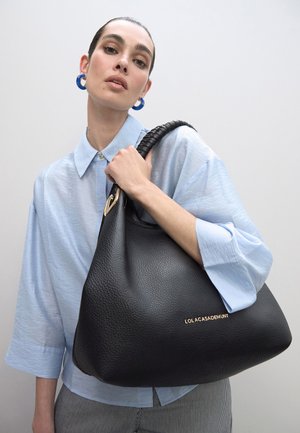 Bolso negro de cuero con acabado texturizado, asa circular grande y acento de herrajes dorados. Camisa azul claro con mangas abullonadas.
