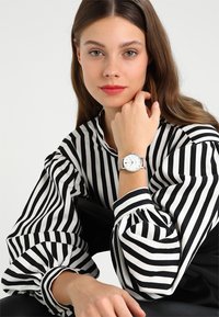 El reloj presenta una caja redonda de color plata, una esfera blanca con números de hora negros minimalistas y una correa gris. El modelo lleva una blusa de rayas en blanco y negro.