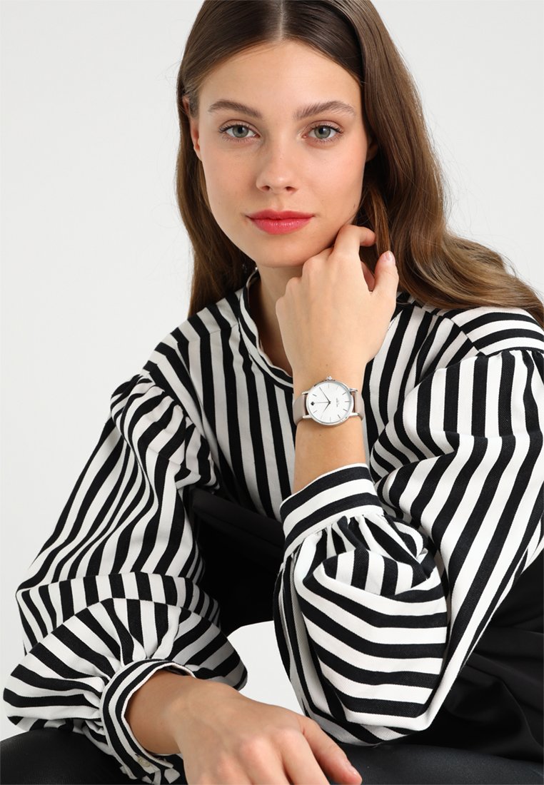El reloj presenta una caja redonda de color plata, una esfera blanca con números de hora negros minimalistas y una correa gris. El modelo lleva una blusa de rayas en blanco y negro.
