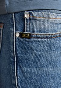 Pantalones vaqueros de mezclilla azul con costuras naranjas, herrajes plateados y una etiqueta negra con la inscripción "Tiger of Sweden" cerca del bolsillo delantero.