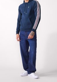 Sudadera marinera con cordón y bolsillo frontal, con franjas rojas y blancas en las mangas. Combinada con jeans azules y zapatillas blancas.