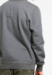 Sudadera gris con cuello redondeado, puños y cinturilla acanalados. Cuenta con un pequeño y sutil bolsillo en la parte posterior y una textura suave.