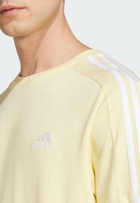 Camiseta de algodón amarilla con acentos de tres franjas blancas en los hombros y un pequeño logo bordado en púrpura claro en el pecho.