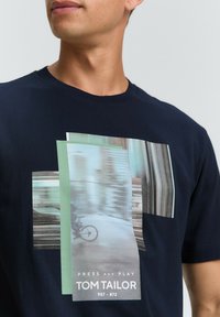 Tricou cu mânecă scurtă de culoare bleumarin, cu imprimeu grafic care prezintă imagini rectangulare suprapuse, text și un motiv cu bicicletă. Fabricat din bumbac.
