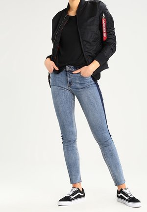 Černá bomber bunda se zipem a červenou nášivkou, kombinovaná s modrými skinny džíny a černými teniskami. Hladká látka, slim fit, ležérní vzhled.
