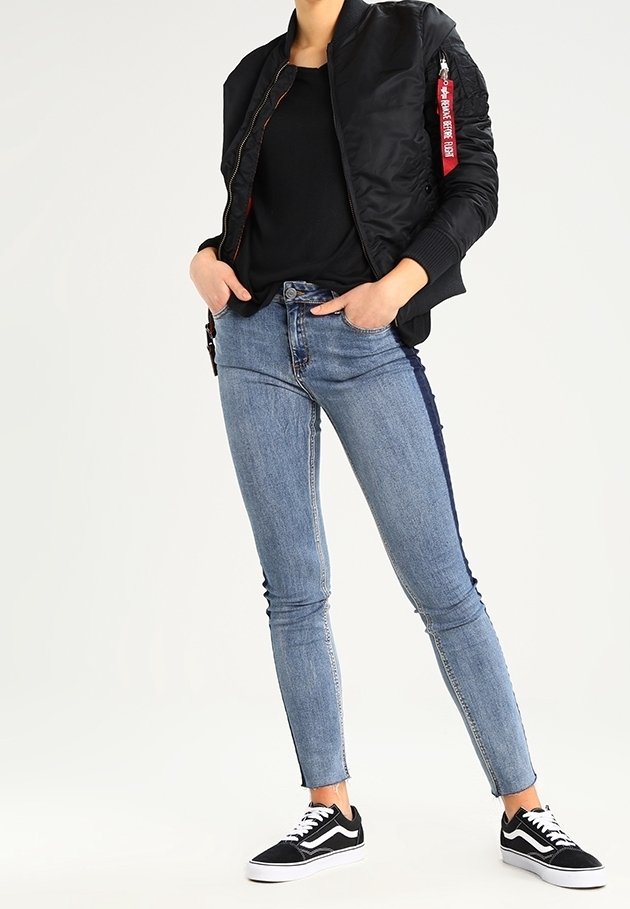 Giubbotto bomber nero con zip e etichetta rossa, abbinato a jeans skinny blu e sneakers nere. Tessuto liscio, vestibilità slim, look casual.