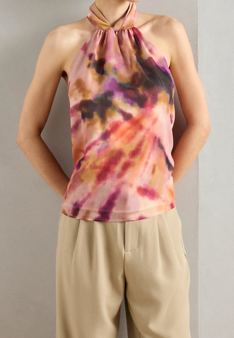 Personne portant un haut sans manches à col noué tie-dye aux teintes rose, orange et marron, associé à un pantalon beige ample.
