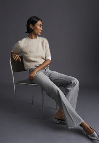 Donna con capelli corti neri che indossa un maglione beige peloso, jeans grigi e tacchi argento, seduta di lato su una sedia di metallo su sfondo grigio.