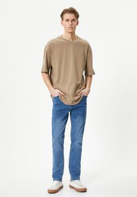 T-shirt beige oversize con maniche corte, abbinata a jeans azzurri chiari e sneakers bianche con suola marrone. Design semplice e texture morbida.