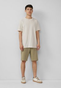Helle beige strukturierte Kurzarm-T-Shirt kombiniert mit olivgrünen Shorts und beigen Sneakers mit braunen Akzenten. Einfaches, lässiges Outfit.