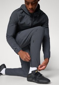 Donkergrijze sportjas met capuchon en twee zakken met rits, gecombineerd met bijpassende grijze broek en zwarte voetbalschoenen met goudkleurige accenten.