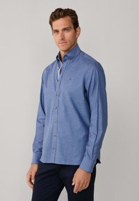 Svijetloplava košulja na kopčanje s raširenim ovratnikom, dugim rukavima i smeđim gumbima. Karakterizira ju diskretna tekstura tkanine i mali logotip na prsima.