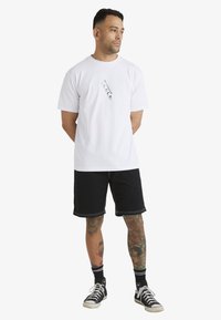 Camiseta blanca de algodón con un estampado gráfico en el frente, combinada con pantalones cortos negros con un detalle de costura en contraste y zapatillas de caña alta.