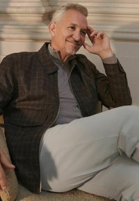 Gray plaid jacket over a gray zip-neck shirt, paired with light gray pants, seated on a textured surface, hand resting on chin.